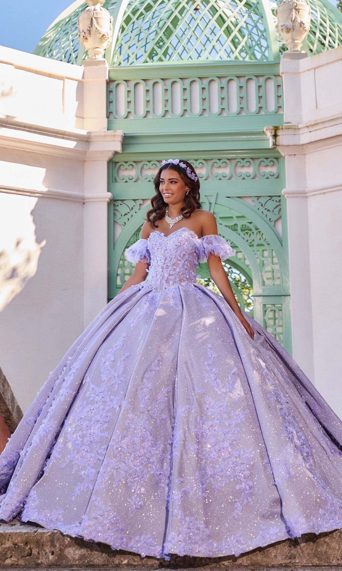 A young woman smiles as she poses before a mint green domed structure, her hands resting gracefully on the ornate lilac Quinceanera Dress PR30134 By Princesa, featuring a strapless corset bodice and voluminous ball gown skirt.